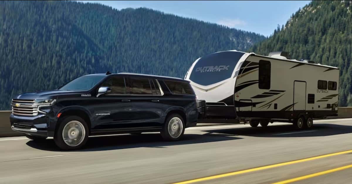 Blue 2023 Chevrolet Suburban Towing An RV Camper Down The Road With A Pine Tree Mountain Side Background