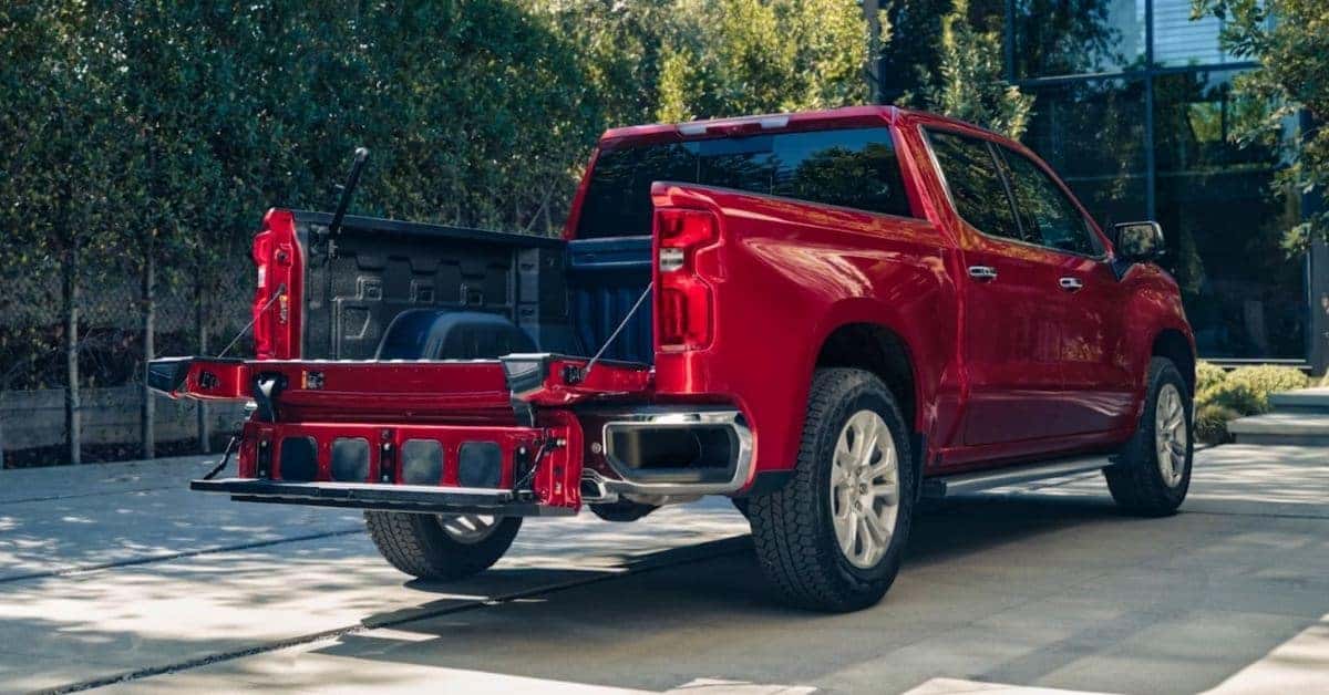 A vibrant red Chevrolet Silverado 1500 pickup truck showcasing its innovative multi-functional tailgate, parked on a paved driveway with lush greenery and a modern building in the background.