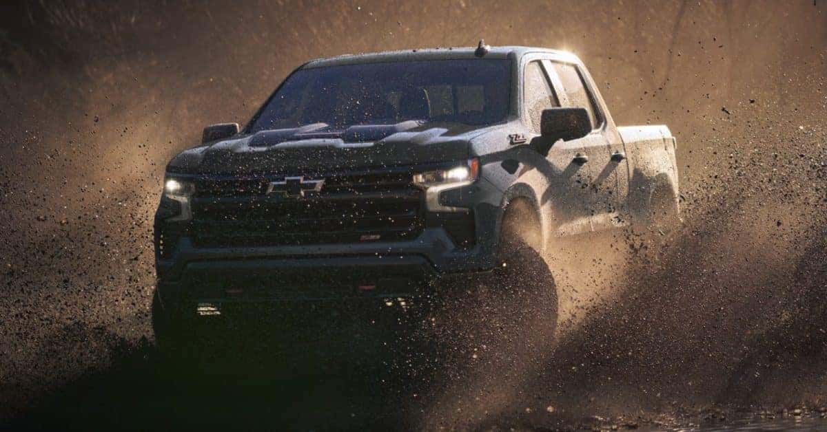 A dark-colored Chevrolet Silverado 1500 pickup truck driving through muddy terrain, creating a dramatic splash of mud and water. The truck's front grille, headlights, and off-road tires are highlighted, with a natural earthy background and golden hour lighting.