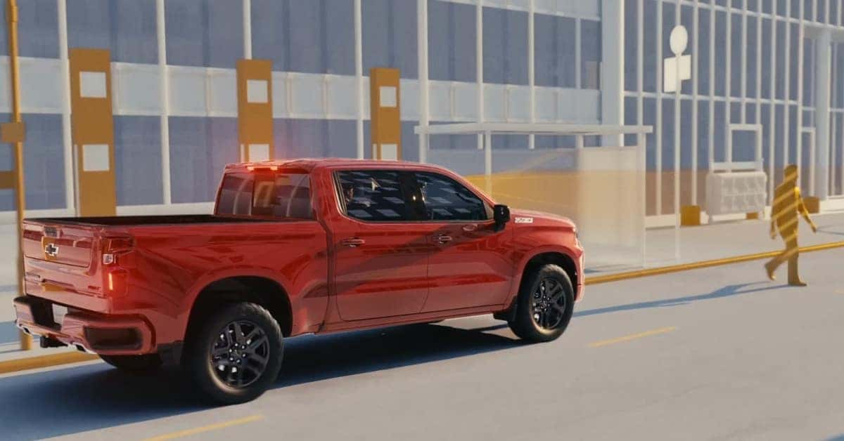 A stylized red Chevrolet Silverado 1500 driving on a paved road next to a modern building, with illuminated taillights and a translucent pedestrian silhouette walking on the sidewalk.