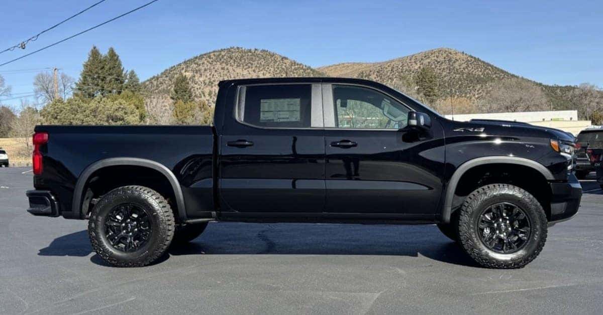 A sleek black Chevrolet Silverado 1500 pickup truck with a Z71 or ZR2 off-road package, parked on an asphalt lot against a backdrop of rolling hills and a clear blue sky. The truck features aggressive all-terrain tires, black fender flares, and a lifted stance.