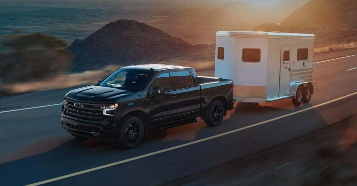 A black Chevrolet Silverado 1500 towing a white horse trailer on a scenic highway at sunrise or sunset, with a mountainous landscape and warm-toned sky in the background.