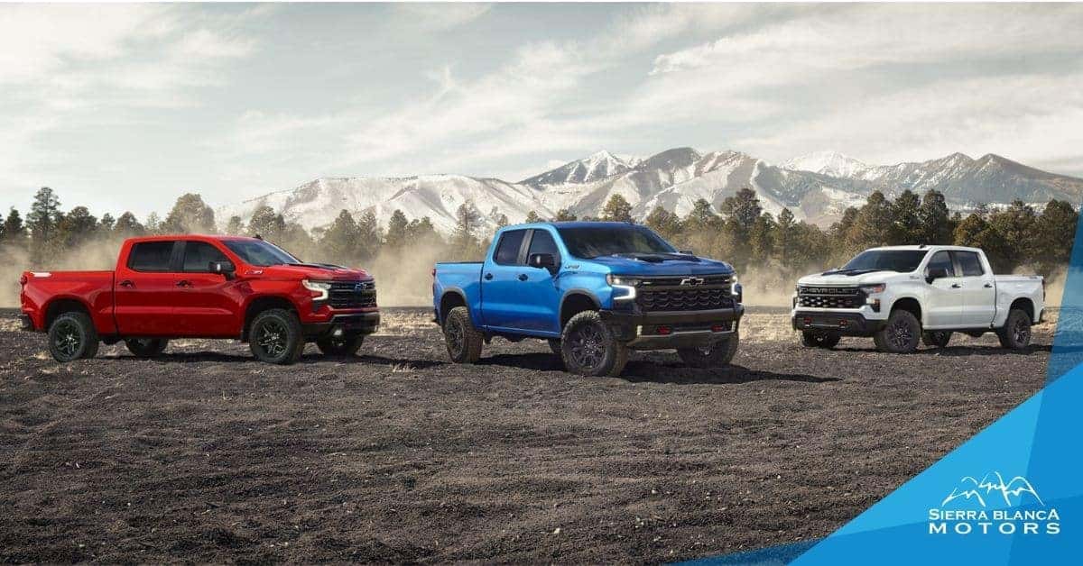 A lineup of three Chevrolet Silverado 1500 trucks in red, blue, and white, parked on rugged terrain with a forest and snow-capped mountains in the background, promoting their off-road capabilities.
