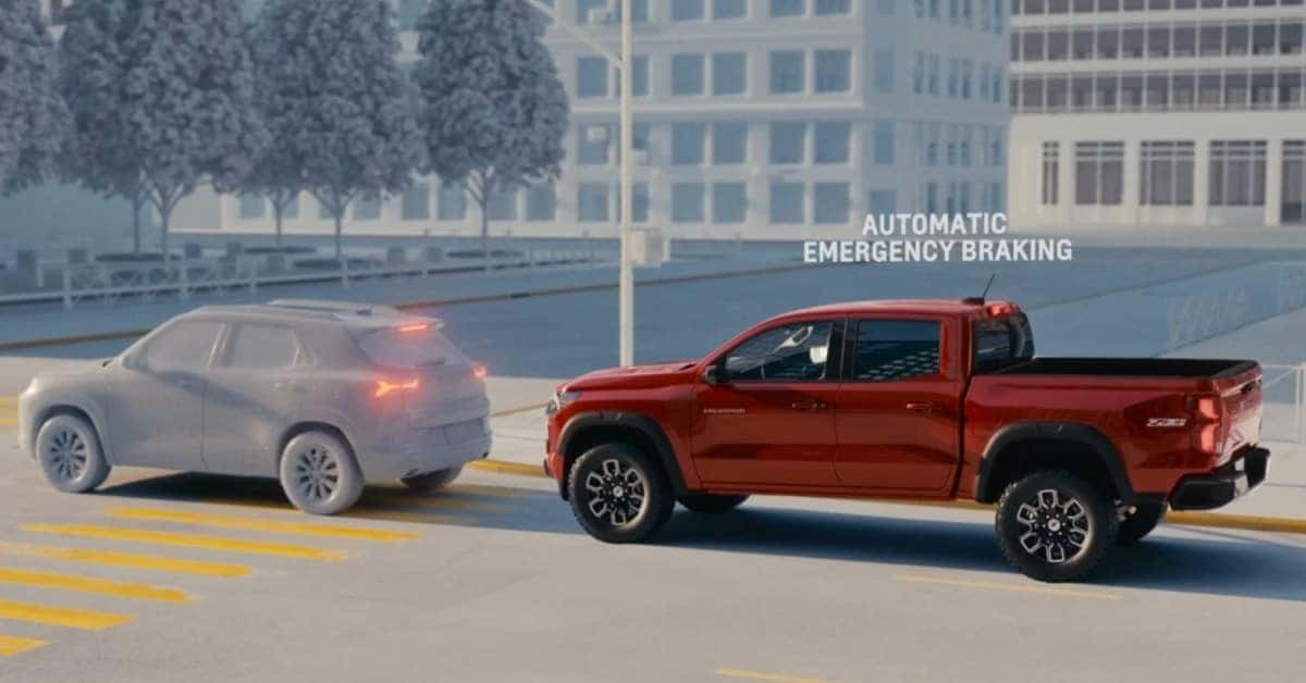 A red 2026 Chevrolet Colorado ZR2 pickup truck demonstrating automatic emergency braking in a city setting. The truck approaches a gray SUV with illuminated brake lights, with text overlay reading 'AUTOMATIC EMERGENCY BRAKING.' The background features modern buildings and trees.