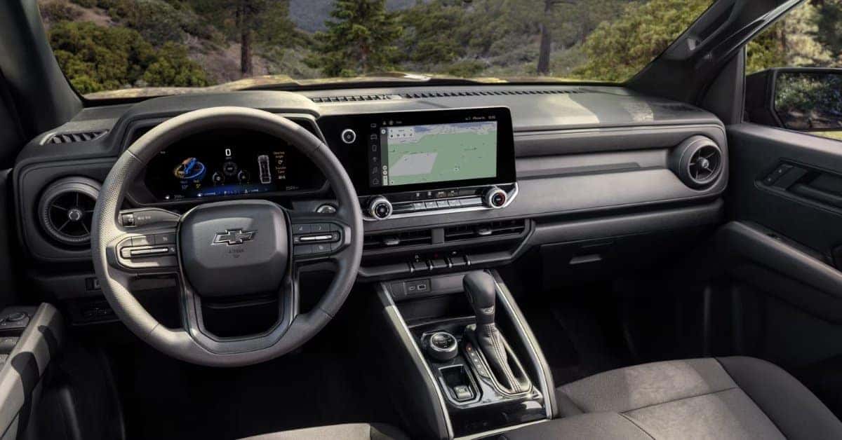 The interior of a 2026 Chevrolet Colorado, highlighting a modern dashboard with a digital instrument cluster and infotainment touchscreen. The steering wheel features the Chevrolet logo, and the windshield reveals a forested outdoor scene.
