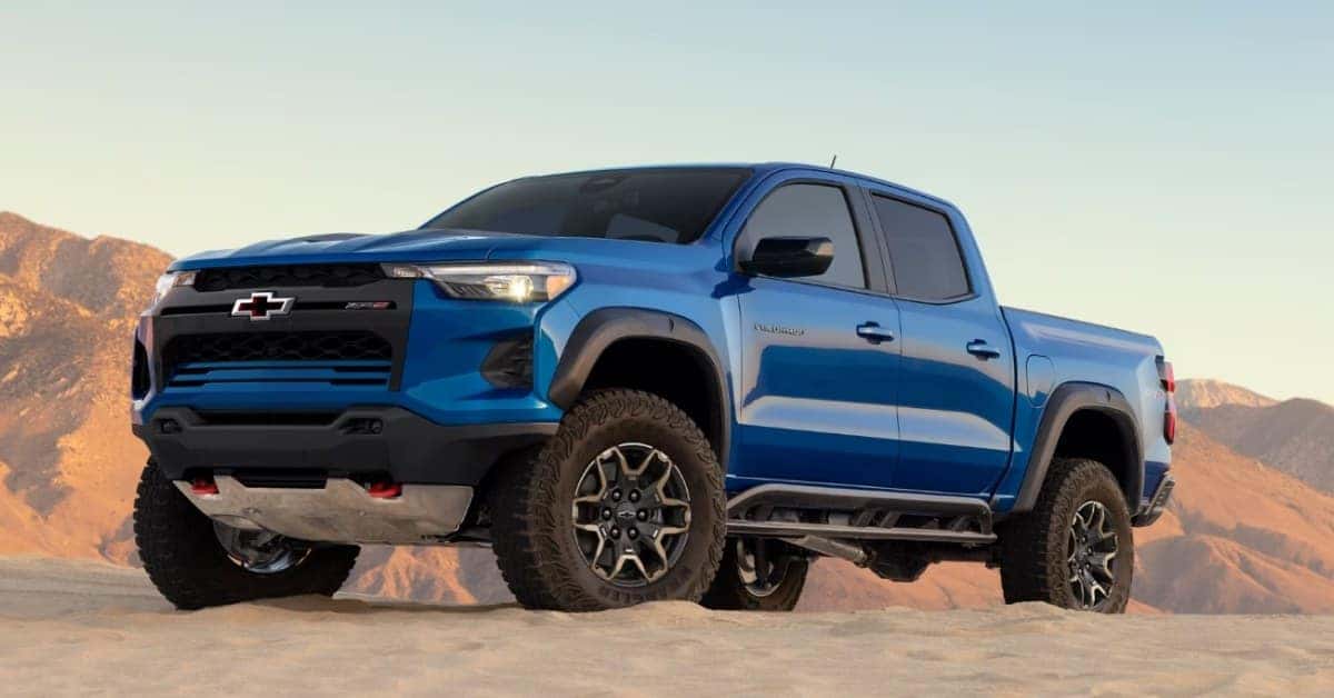 A vibrant blue 2026 Chevrolet Colorado ZR2 pickup truck parked on sandy, desert-like terrain with rugged mountains in the background under a clear blue sky. The truck features aggressive off-road tires, black fender flares, and red tow hooks, emphasizing its rugged design and off-road capabilities.