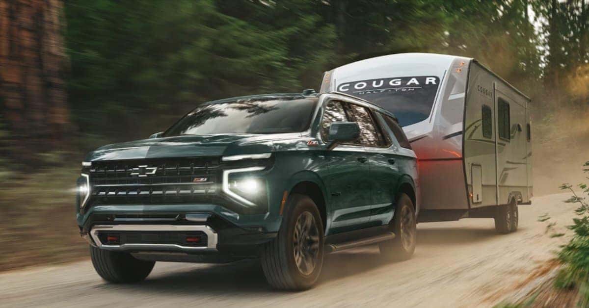 A dark green 2026 Chevrolet Tahoe Z71 SUV towing a white and grey Cougar Half-Ton travel trailer on a dusty, tree-lined dirt road. The SUV features Z71 badging, C-shaped LED headlights, and red tow hooks. Dust trails behind the vehicles, emphasizing motion and adventure. The forest setting highlights the SUV's towing and off-road capabilities.