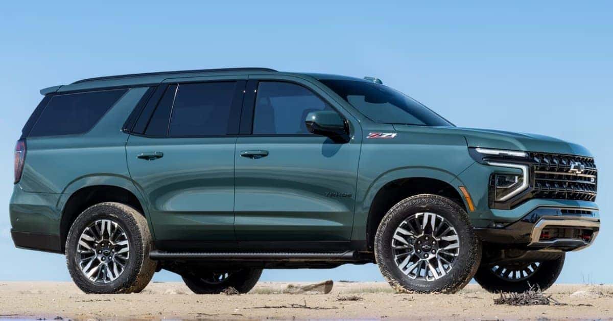 A dark green 2026 Chevrolet Tahoe Z71 SUV parked on a sandy or dirt surface under a clear blue sky. The vehicle features rugged all-terrain tires, Z71 badging, a bold grille with a Chevrolet emblem, and roof rails. The natural setting emphasizes the SUV's off-road readiness and adventurous design.