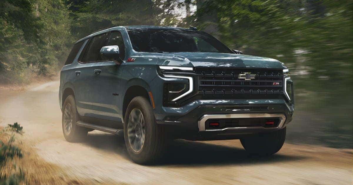A dark green 2026 Chevrolet Tahoe Z71 SUV driving on a dusty, unpaved road through a forest. The vehicle features a bold grille, C-shaped LED headlights, red tow hooks, and rugged tires. Dust is kicked up behind the SUV, emphasizing its off-road capability. The forest background is blurred, creating a dynamic and adventurous atmosphere.