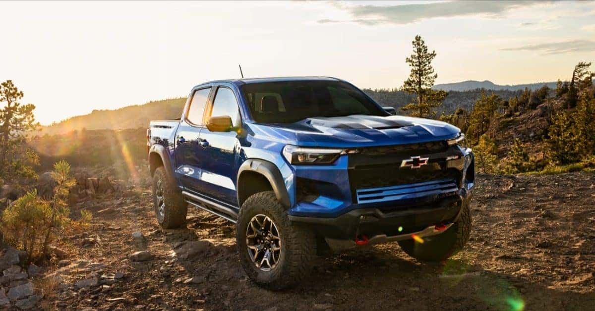 A blue Chevrolet Colorado pickup truck parked on a rocky dirt road in a mountainous environment. The truck features off-road tires, red recovery hooks, and a Chevrolet logo on the grille. The scene is bathed in golden sunlight with a backdrop of evergreen trees and hills.