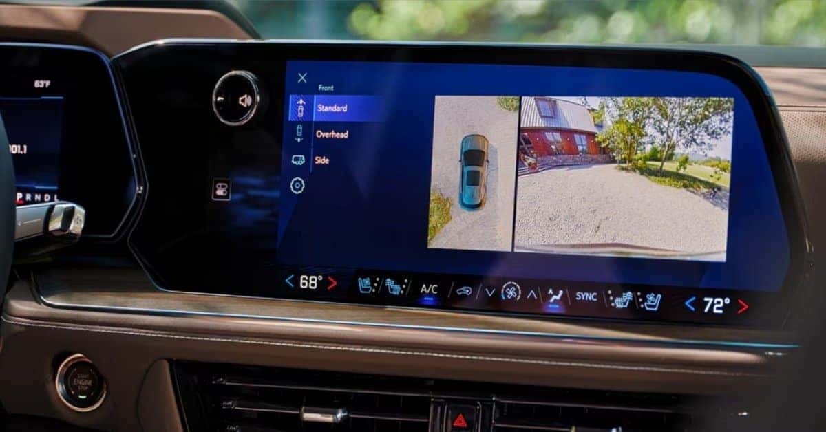 A close-up of a modern 2026 Chevrolet Tahoe's dashboard showcasing a large digital infotainment screen with a 360-degree camera system. The screen displays overhead and wide-angle views of the vehicle's surroundings. Climate controls, including heated and ventilated seat options, are integrated into the display. The dashboard features premium materials, a start/stop engine button, and a sleek, luxurious design.