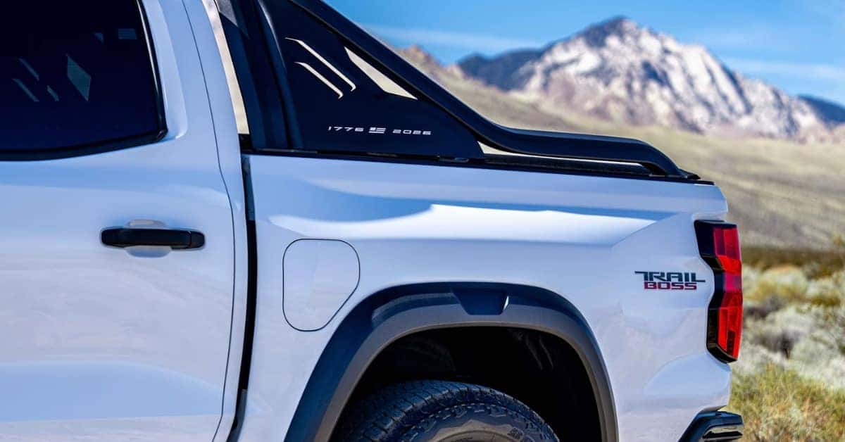 A white 2026 Chevrolet Colorado pickup truck with black accents, showcasing its rear passenger side and bed. The truck features '1776' and '2026' graphics on the rear window trim and a 'TRAIL BOSS' emblem near the tail light. The background includes a mountainous desert landscape under a clear blue sky.