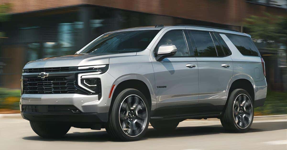 A silver 2026 Chevrolet Tahoe SUV driving on a suburban street, captured from a low-angle front-left perspective. The vehicle features a bold grille with the Chevrolet logo, L-shaped LED headlights, and large alloy wheels in motion. The background is blurred, showcasing urban architecture and greenery, emphasizing the SUV's dynamic presence and modern design.