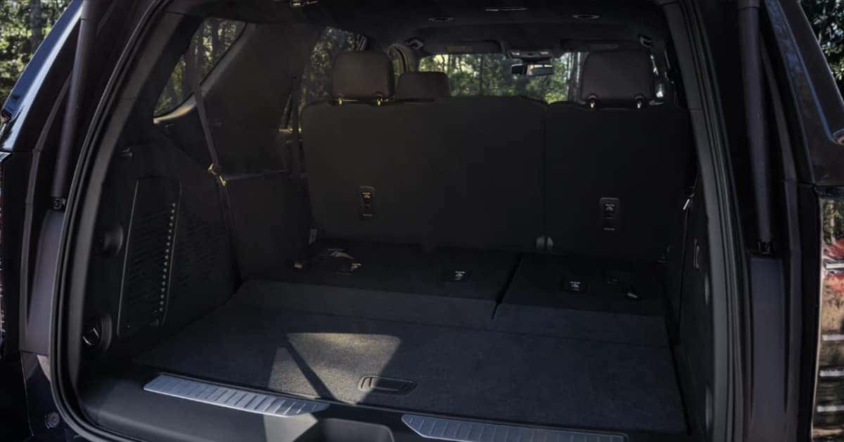The rear cargo area of a dark-colored 2026 Chevrolet Tahoe with the second and third-row seats folded flat, creating a large, continuous storage space. The interior features dark carpeting, seat release mechanisms, and a metallic threshold plate. The background through the windows shows green trees, suggesting the vehicle is parked outdoors.