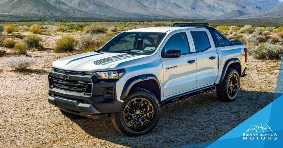 A white 2026 Chevrolet Colorado pickup truck with black accents, parked on a dirt surface in a desert setting. The truck features a black grille, off-road tires, and a roll bar. The background includes rugged mountains under a clear sky, with branding for 'Sierra Blanca Motors' visible in the corner.