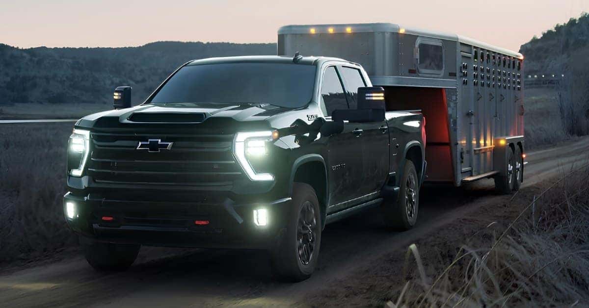 A Chevrolet Silverado 2500 HD Z71 drives on a dirt road at dusk while towing a large livestock trailer through a rural landscape.