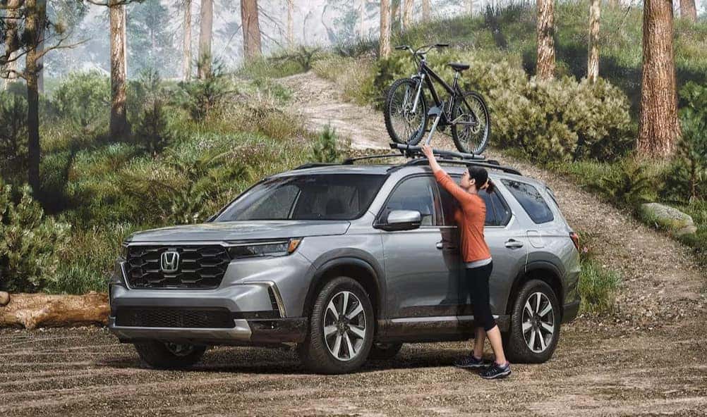 A grey 2025 Honda Pilot sits while the driver loads a bike on top, making this the perfect SUV for on or off-road adventures near Spartanburg.