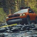 An orange Honda Passport driving off-road by a rocky shallow river