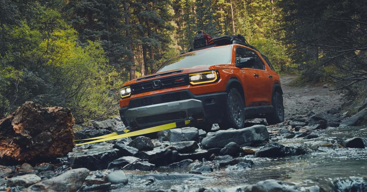 An orange Honda Passport driving off-road by a rocky shallow river