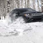 A black 2016 Jeep Compass drives through deep snow in a forest of birch trees