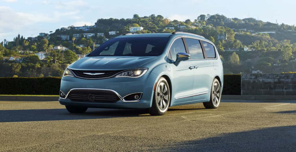 A light blue 2017 Chrysler Pacifica Hybrid parked in front of a wooded hill with white houses