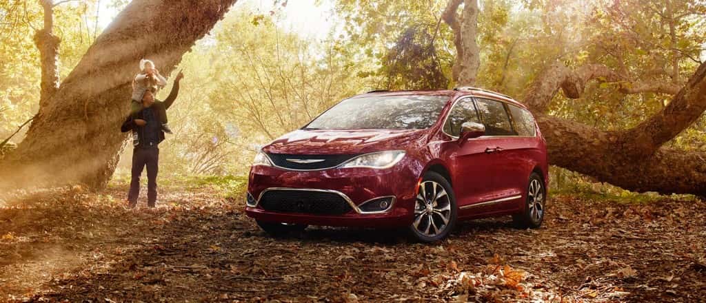 In a sunny forest, a man points to the trees with his daughter on his shoulders as they stand next to their red Chrysler Pacifica. 