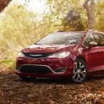 In a sunny forest, a man points to the trees with his daughter on his shoulders as they stand next to their red Chrysler Pacifica.
