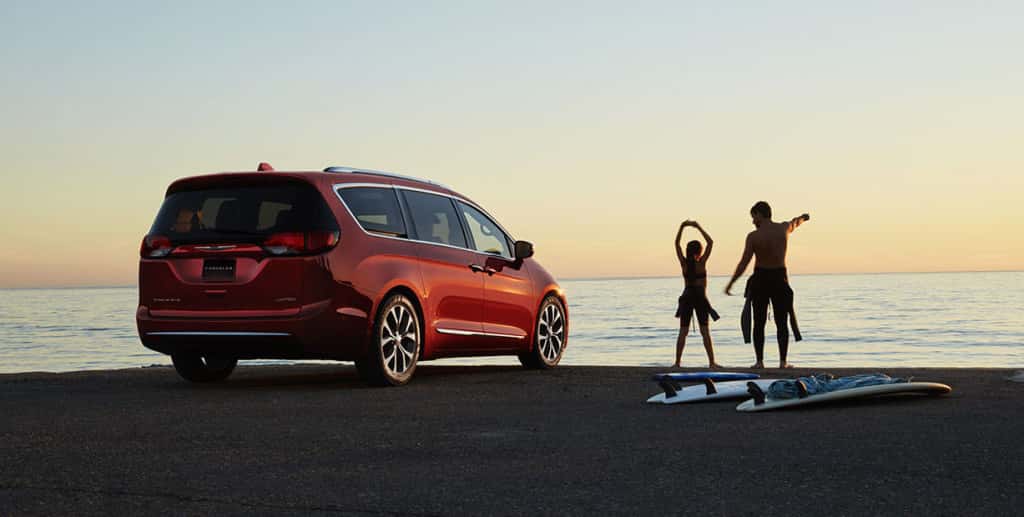 Sillhouetted against the sky and water, two surfers on a beach stand next to a red 2017 Chrysler Pacifica