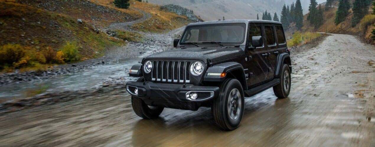 A popular used Jeep Wrangler for sale, a black 2018 Jeep Wrangler Unlimited, is shown driving on a muddy road.