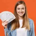 A young blonde woman smiling in a denim button-up and white t-shirt holding up a fan of hundred dollar bills in front of an orange background