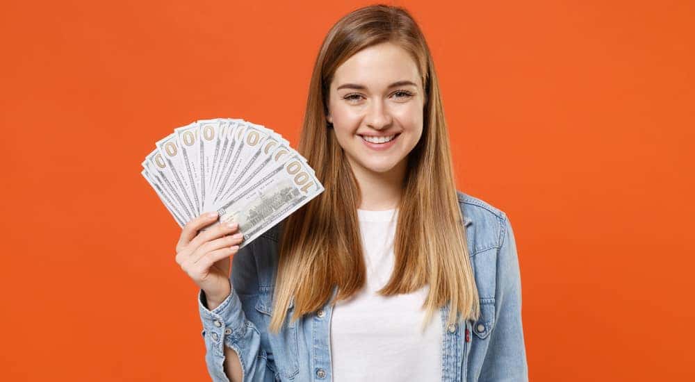 A young blonde woman smiling in a denim button-up and white t-shirt holding up a fan of hundred dollar bills in front of an orange background