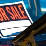 Close-up of an orange, black, and white "FOR SALE" sign in the front window of a vehicle with the hood open
