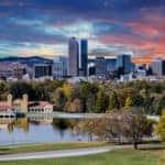 Green grass, trees, and a pond in front of tall Denver buildings, mountains, and an orange sky