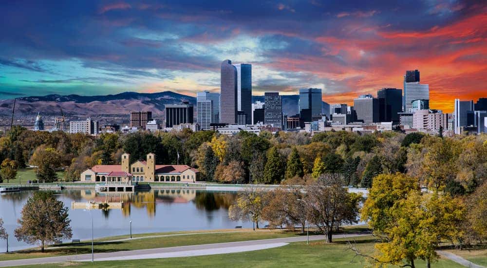 Green grass, trees, and a pond in front of tall Denver buildings, mountains, and an orange sky
