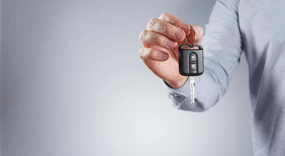 Abdomen, arm, and hand of man in a grey shirt holding up a black car key in front of a grey background