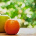 Green apple and an orange sitting on a brown wooden table in front of green plants