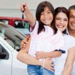 Mom, dad, son, and daughter holding a car key, all standing near cars in a dealership showroom