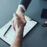 Handshake over a grey table with a clipboard, phone, and another hand holding a car key on it