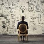 Man sitting in a brown chair facing a wall covered in writing and drawings that indicate planning and strategy