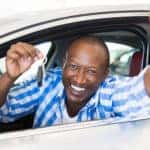 Man in a blue and white plaid shirt sticking his head and arms out the window of a white vehicle, holding out car keys, and smiling
