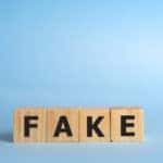 Four brown wooden blocks with black letters on them that spell out the word "FAKE" against a blue background