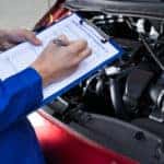 Man in a blue shirt, writing on a clipboard above the open hood of a red car