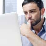 Man with facial hair in a blue shirt sitting at a desk with a laptop