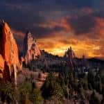 Tall orange rock mountains are shown surrounded by trees in front of an orange, cloudy sky.