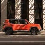 An orange 2019 Jeep Renegade is parked outside a building on a shady street. Check one out at a Jeep dealership in Colorado Springs.