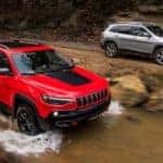 A red 2019 Jeep Cherokee is driving in a river while a grey one drives on the trail.