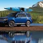 A blue 2010 Ford Escape is parked between a river and a mountain with a kayak on the roof.