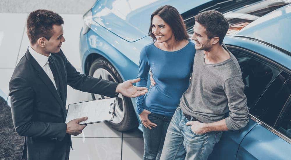 A salesman is explaining to a couple leaning on a blue car why Certified Pre-Owned cars are a good choice.