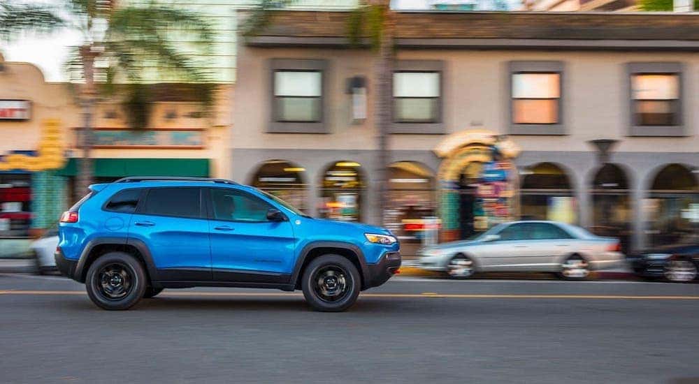 A blue 2019 used Jeep Cherokee is driving on a city street and is shown from the passenger side.