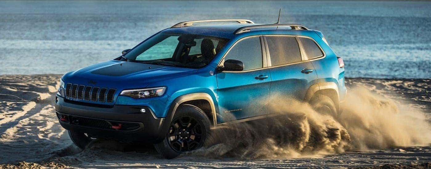 A blue 2019 used Jeep Cherokee Trailhawk is kicking up sand on a beach.